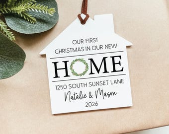 May include: A white house-shaped ornament with a brown string, featuring the text OUR FIRST CHRISTMAS IN OUR NEW HOME, with a green wreath replacing the letter O. It includes the address 1250 SOUTH SUNSET LANE, the names Natalie & Mason, and 2026.