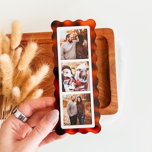 Peut inclure: Un cadre photo rectangulaire avec un motif écaille de tortue et un bord ondulé. Le cadre contient trois photos verticales de personnes et de chiens. Le cadre est tenu dans la main d'une personne, avec un bouquet de fleurs séchées en arrière-plan.