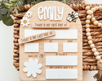 May include: A wooden sign with a bee and daisy design. The sign is personalized with the name "Emily" and has spaces to fill in information about the child's first and last day of school, age, height, things they love, their teacher's name, and what they want to be when they grow up.
