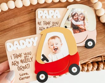 May include: Wooden photo frames in the shape of toy cars. One is red and yellow with the text "DADDY" and "I THINK YOU ARE WHEELY GREAT!" The other is pink and yellow with the text "PAPA" and "WE THINK YOU ARE".