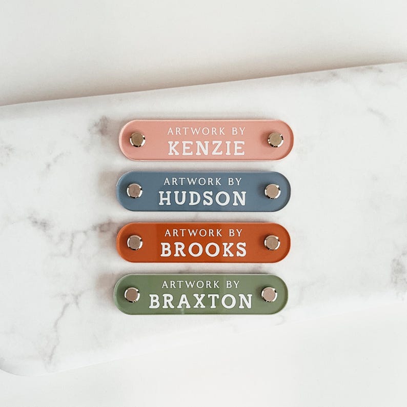 K&ouml;nnte beinhalten: Vier farbige Namensschilder mit dem Text "Artwork by" gefolgt von einem Namen. Die Schilder sind rosa, blau, orange und gr&uuml;n, jedes mit silberner Hardware. Die Namen sind Kenzie, Hudson, Brooks und Braxton.