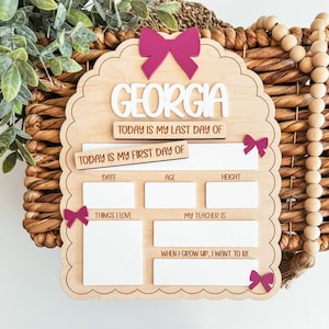 May include: A wooden school memory board with the name "GEORGIA" in white. The board includes spaces for "TODAY IS MY LAST DAY OF," "TODAY IS MY FIRST DAY OF," date, age, height, things I love, teacher, and aspirations. It has pink bow accents.