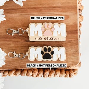 Puede incluir: Dos llaveros con la palabra "MOM" en letras blancas, con una huella de pata en el centro. Un llavero es de color rubor con una huella de pata rosa y los nombres "milo & lillian". El otro es negro con una huella de pata negra.