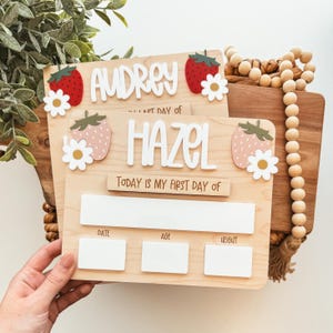 May include: Wooden milestone boards with the names "Audrey" and "Hazel" in white lettering. Each board features strawberry and daisy accents. The bottom section has spaces for date, age, and weight.