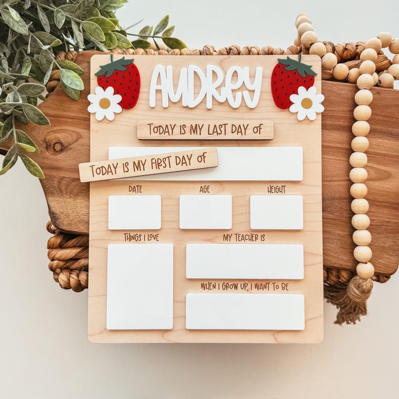 May include: A wooden milestone board with the name "Audrey" in white letters, adorned with strawberry and daisy accents. The board includes sections for "Date," "Age," "Height," "Things I Love," "My Teacher Is," and "When I Grow Up, I Want to Be."