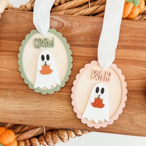 May include: Two Halloween-themed wooden ornaments. Each ornament features a white ghost holding an orange pumpkin. One has a green scalloped border and the word "CREW", the other has a pink scalloped border and the word "RYK". Both have white ribbon.