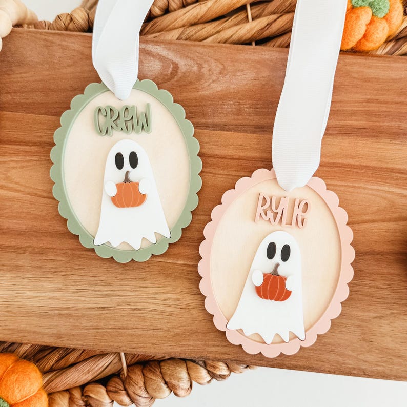 May include: Two Halloween-themed wooden ornaments. Each ornament features a white ghost holding an orange pumpkin. One has a green scalloped border and the word "CREW", the other has a pink scalloped border and the word "RYK". Both have white ribbon.