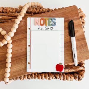 May include: White dry-erase board with the word "NOTES" in colourful letters and the name "Ms. Smith". A red apple and a black dry-erase marker are also visible. The board is on a wooden cutting board.