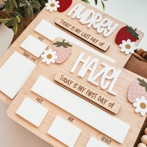 May include: Two wooden milestone boards with the names "Audrey" and "Hazel" in white script. Each board has space for date, age, and height, with strawberry and daisy accents. Text reads "Today is my last day of" and "Today is my first day of".