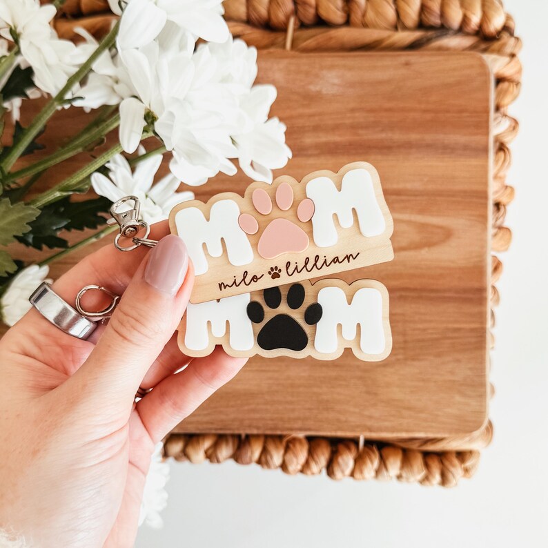 Puede incluir: Dos llaveros de madera con la palabra "MOM" en letras blancas y huellas de patas. Un llavero tiene una huella de pata rosa, el otro una huella de pata negra. Los nombres "milo & lillian" est&aacute;n grabados en el llavero superior.
