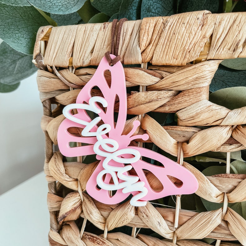 May include: Pink butterfly-shaped ornament with the word "bloom" written in white on it. The ornament is hanging from a brown string and is in front of a woven basket.