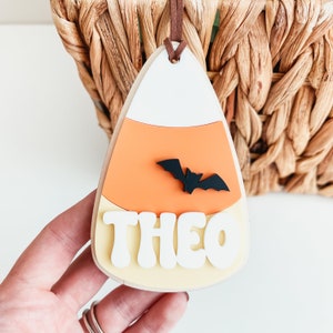 May include: A wooden candy corn shaped ornament with a black bat and the name "THEO" in white letters.