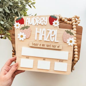 May include: A wooden milestone board with the names "Audrey" and "Hazel" in white letters. The board features strawberry and daisy accents. Text reads "Today is my first day of" with spaces for date, age, and weight.