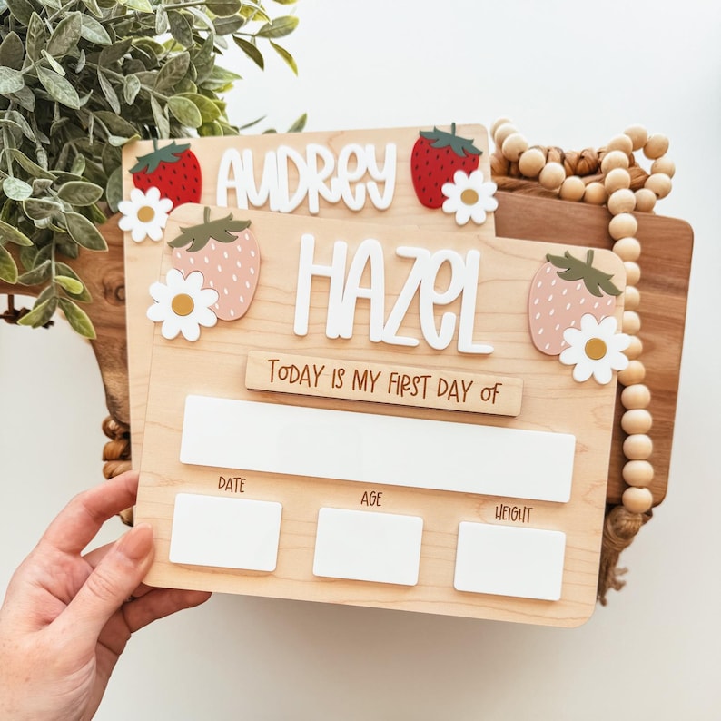 May include: A wooden milestone board with the names "Audrey" and "Hazel" in white letters. The board features strawberry and daisy accents. Text reads "Today is my first day of" with spaces for date, age, and weight.