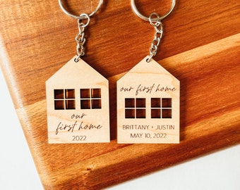 May include: Two wooden house-shaped keychains with silver key rings. One keychain says "our first home 2022" and the other says "our first home BRITTANY + JUSTIN MAY 10, 2022".