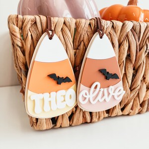 May include: Two Halloween-themed candy corn ornaments with bats and names "Theo" and "Olive" written in white. The ornaments are hanging from a wicker basket.