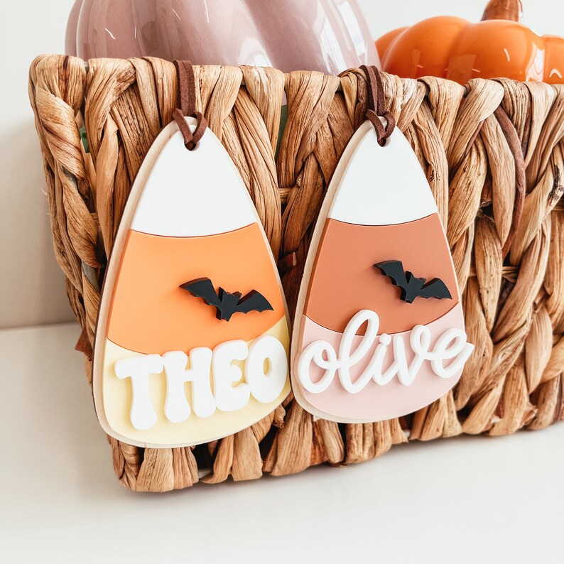 May include: Two Halloween-themed candy corn ornaments with bats and names "Theo" and "Olive" written in white. The ornaments are hanging from a wicker basket.