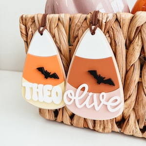 May include: Two candy corn shaped ornaments with the names Theo and Olive written in white on them. Each ornament has a black bat silhouette on the orange section.