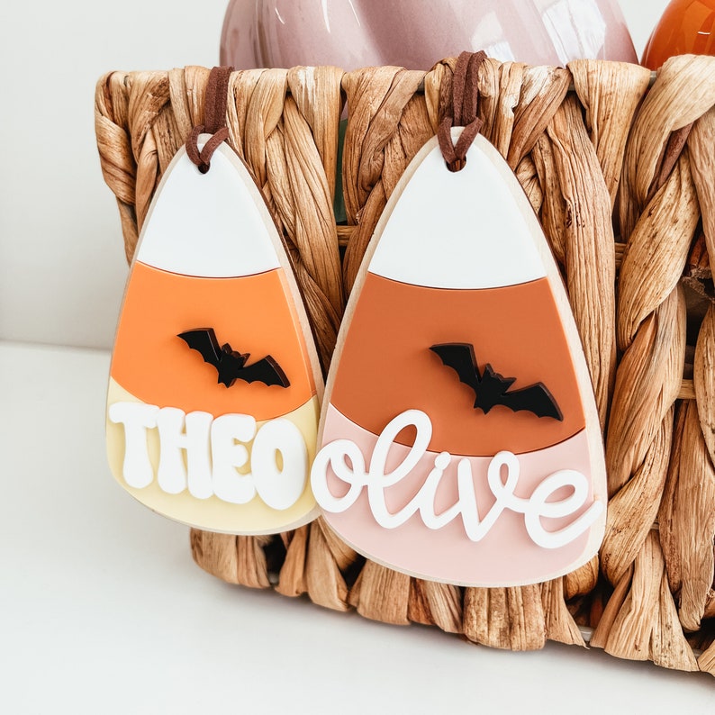 May include: Two candy corn shaped ornaments with the names Theo and Olive written in white on them. Each ornament has a black bat silhouette on the orange section.