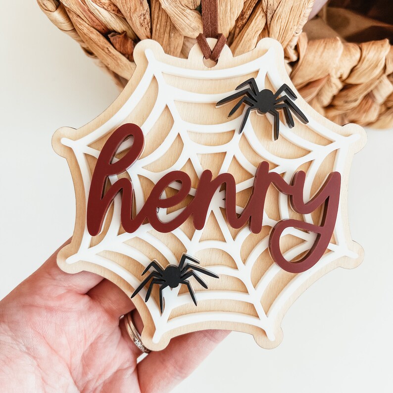 May include: A white wooden spiderweb ornament with the name "Henry" in maroon lettering and two black spiders.