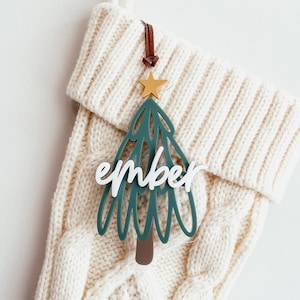 Peut inclure: Une chaussette en tricot blanche avec une décoration de sapin de Noël verte et dorée qui dit "ember" dessus.