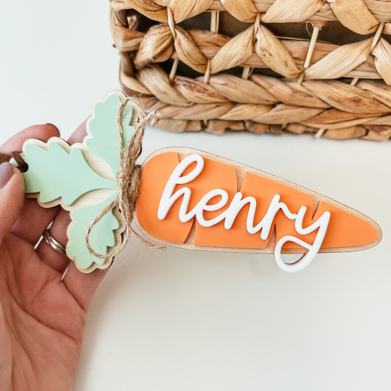 May include: A wooden carrot-shaped ornament with a green leafy top and the name "Henry" written in white on the orange carrot part.