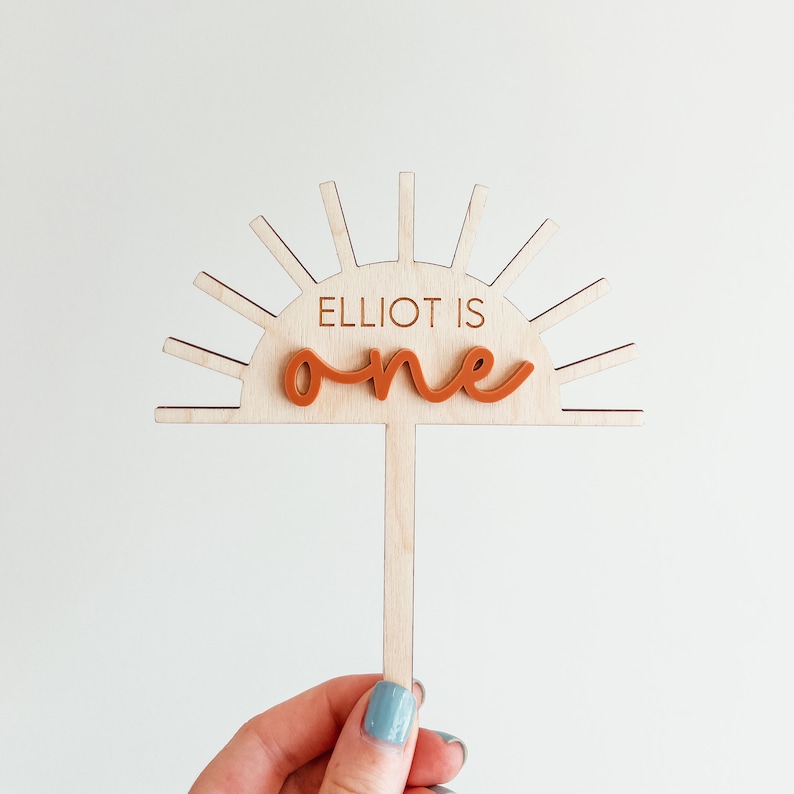 Puede incluir: Topper de pastel de madera con forma de sol con el texto "Elliot is one" en letras marrones. El sol tiene un palo de madera unido a la parte inferior.