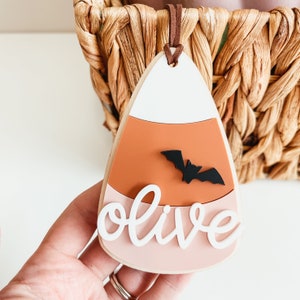 May include: A white, orange, and pink candy corn shaped ornament with a black bat and the name "olive" written in white.