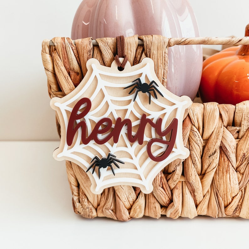 May include: A white wooden spiderweb ornament with the name "Henry" written in maroon letters. The ornament has two black spiders on it.