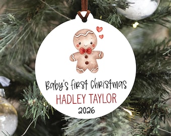 May include: A round white Christmas ornament featuring a watercolor gingerbread person with a red bow tie and two small red hearts. The text reads Baby's first Christmas, HADLEY TAYLOR, and 2026. A brown leather cord is attached to the top.
