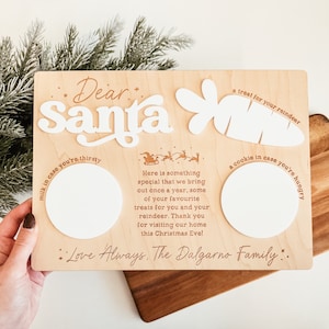 May include: A wooden sign with the words "Dear Santa" in white letters. The sign has three white circles for milk, a cookie, and a carrot treat for Santa's reindeer. The sign also includes the text "Love Always. The Dalgarno Family."