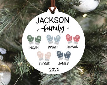 May include: A round white Christmas ornament featuring the text JACKSON family at the top and 2026 at the bottom. It displays five pairs of illustrated mittens in green, light blue, pink, beige, and dark blue, each labeled with names Noah, Wyatt, Rowan, Elodie, and James.