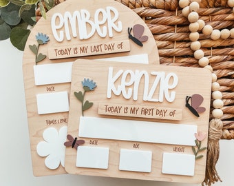 May include: Two wooden signs with a floral and butterfly theme. The top sign says "Ember" and "Today is my last day of". The bottom sign says "Kenzie" and "Today is my first day of". Both signs have spaces for writing the date, age, height, and things they love.