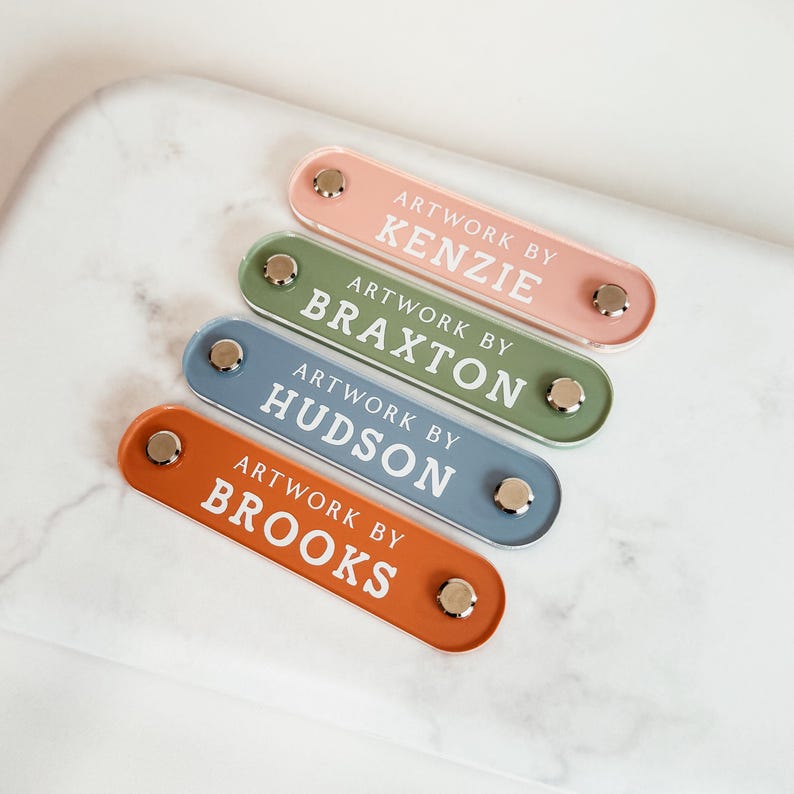 K&ouml;nnte beinhalten: Vier farbige Namensschilder mit dem Text "ARTWORK BY" und den individuellen Namen: Kenzie, Braxton, Hudson und Brooks. Die Schilder sind in den Farbt&ouml;nen Rosa, Gr&uuml;n, Blau und Orange gehalten und haben silberfarbene Beschl&auml;ge.