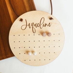 May include: A round wooden earring holder with the name "Jacqueline" engraved on it. The holder has multiple holes for storing earrings. Two pairs of earrings are displayed on the holder, one pair is pink and sparkly, the other pair is white and pearlescent.