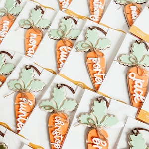 May include: A set of wooden carrot ornaments with different names written on them. The carrots are painted orange and have green tops. The names are written in white paint.