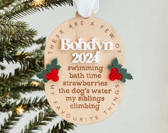 May include: A wooden Christmas ornament with the text "THESE ARE A FEW OF MY FAVORITE THINGS" surrounding a circle with the text "Bohdyn 2024" and a list of favorite things: swimming, bath time, strawberries, the dog's water, my siblings, climbing.
