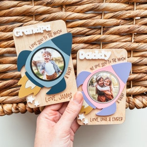 Peut inclure: Deux cartes en bois en forme de fusée avec des photos et des messages personnalisés. La carte de la fusée bleue dit "Grandpa I love you to the moon & back! Love, James". La carte de la fusée rose dit "Daddy We love you to the moon & back! Love, Luna & Ember".