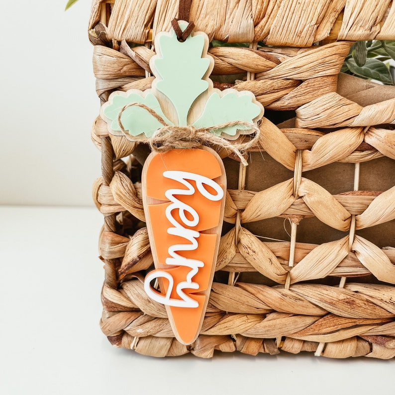 May include: A wooden carrot-shaped ornament with a green leafy top and the name "Henry" written in white on the orange carrot. The ornament is hanging from a woven basket.