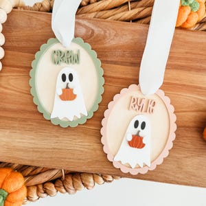 May include: Two Halloween-themed wooden ornaments. Each ornament features a scalloped edge, a light-colored center, and a ghost holding an orange pumpkin. One says "CREW" and the other says "RUR". Both have white ribbons.