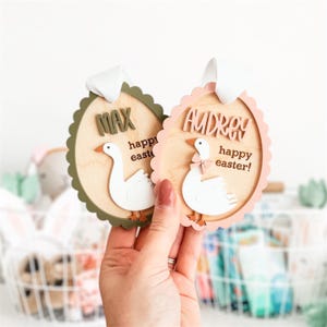 May include: Two wooden Easter ornaments shaped like eggs with a goose design. The ornaments are decorated with the names "Max" and "Audrey" and the text "happy easter!" in pink and green paint.
