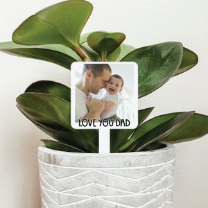 May include: White plant marker with a photo of a father and child smiling. The text "LOVE YOU DAD" is printed below the photo.