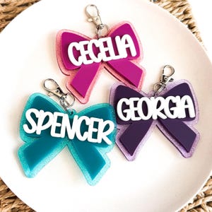May include: Three personalized bow-shaped keychains in shades of teal, purple, and magenta. Each keychain features a name in white block letters: "Cecelia", "Spencer", and "Georgia". The keychains have silver-colored clasps.