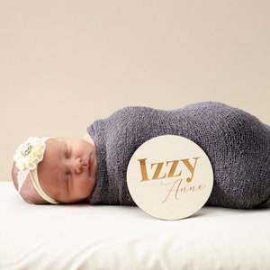 May include: A newborn baby girl is sleeping on a white blanket. She is wearing a white headband with a flower and a gray swaddle. A wooden round sign with the name "Izzy Anne" is next to her.