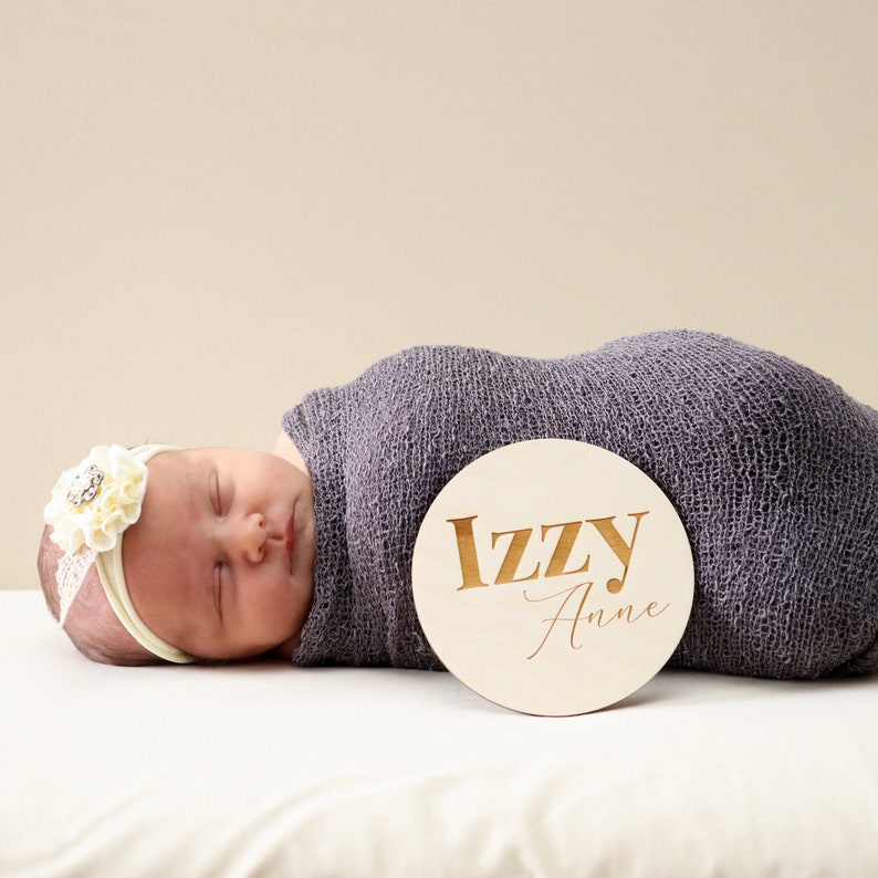 May include: A newborn baby girl is sleeping on a white blanket. She is wearing a white headband with a flower and a gray swaddle. A wooden round sign with the name "Izzy Anne" is next to her.