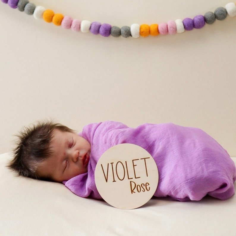 May include: A newborn baby girl is swaddled in a purple blanket and is sleeping next to a wooden circle with the name "VIOLET Rose" written on it.