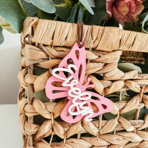 May include: Pink butterfly-shaped wooden ornament with the word "Welcome" written in white on it. The ornament is hanging from a brown string and is in front of a wicker basket.