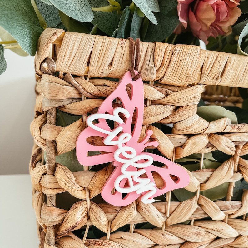 May include: Pink butterfly-shaped wooden ornament with the word "Welcome" written in white on it. The ornament is hanging from a brown string and is in front of a wicker basket.