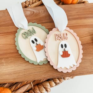 May include: Two round wooden Halloween ornaments with scalloped edges. One is green and the other is pink. Each features a white ghost holding an orange pumpkin and the names "CREW" and "RYLIE" in wooden letters.