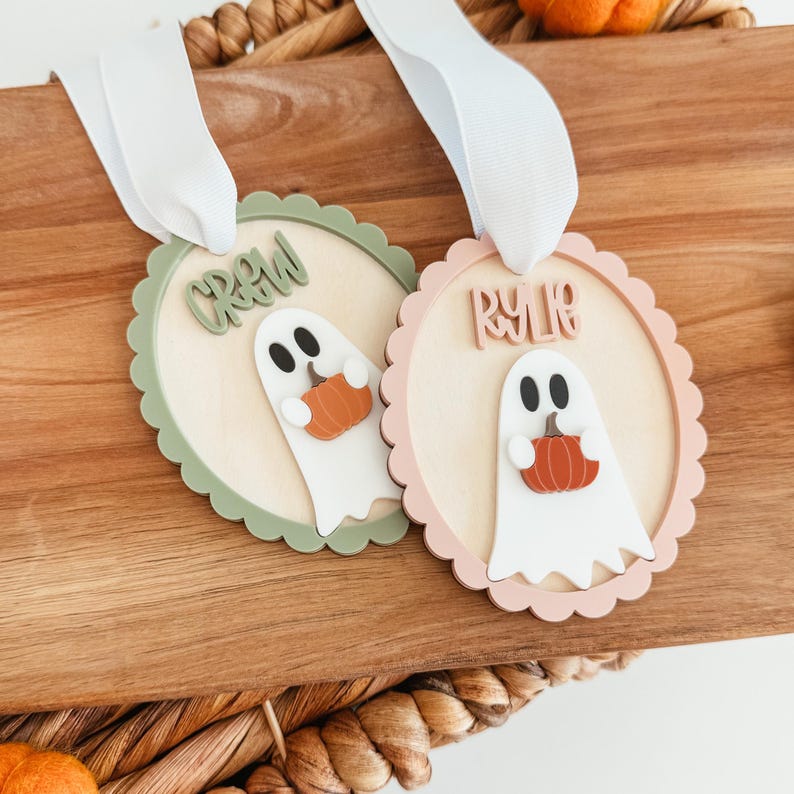 May include: Two round wooden Halloween ornaments with scalloped edges. One is green and the other is pink. Each features a white ghost holding an orange pumpkin and the names "CREW" and "RYLIE" in wooden letters.